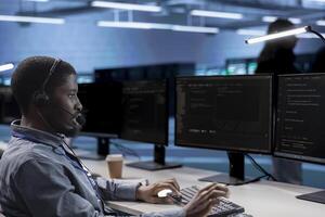 Employee working in call center, providing outsourcing services for data center businesses. African american man at work talking in headphones microphone with on field technicians in server hub photo