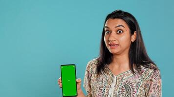 Tech content creator doing influencer marketing using green screen smartphone, isolated over studio background. Indian woman holding empty copy space mockup phone, doing review, camera B photo