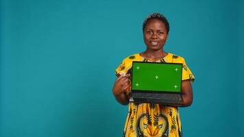 Young adult showing a laptop with isolated mockup screen in studio, presenting a display with green screen against blue background. Female model posing with chroma key. Camera B. photo