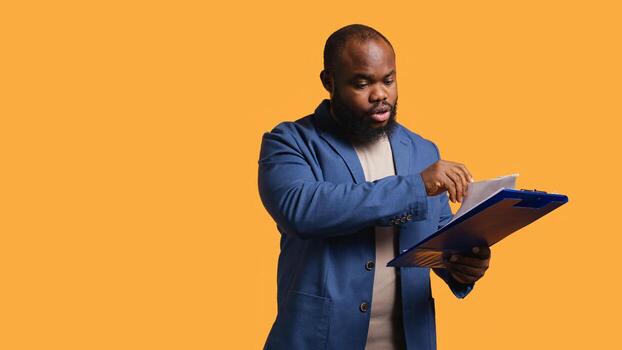 Man browsing though financial audit report on clipboard, pleased by high income, studio background. Happy african american worker reading paperwork on notepad, satisfied by figures, camera B photo