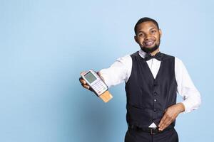 Professional hotel porter presenting the pos terminal and credit card, symbolizing the payment at front desk against blue background. Receptionist with tie asking for the transaction. photo