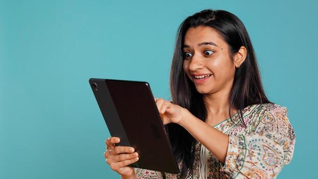 Joyous woman excited to purchase products, adding payment method on website, isolated over studio background. Happy customer does internet shopping, typing debit card info on tablet, camera B photo