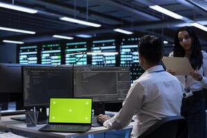 Teamworking technicians in server room using green screen laptop to find network issues affecting supercomputers. IT experts fixing slow data transfer rates in data center with chroma key notebook photo