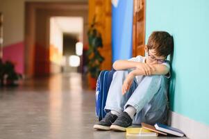Little boy sitting alone on floor after suffering an act of bullying. Sad young schoolboy sitting on corridor with hands on knees and head between his legs photo