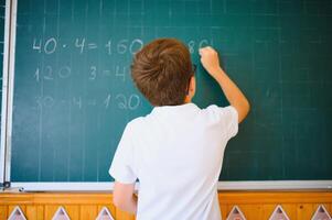 Cute little schoolboy diligent write chalk solve math problem dressed stylish uniform classroom blackboard background photo