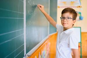 Cute little schoolboy diligent write chalk solve math problem dressed stylish uniform classroom blackboard background photo
