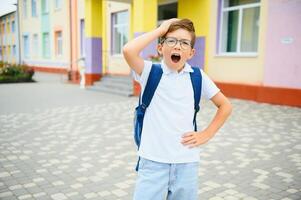 Portrait of little schoolboy in glasses and with backpacks standing on the school building. Study concept photo