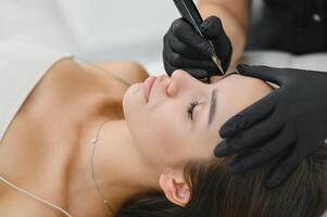 Beauty masters hands do permanent eyebrow makeup. Eyebrow microblading is performed using manipulator handle and special nozzle with needles photo