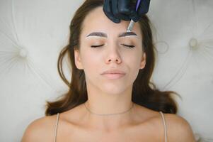 The master holds a white pencil and draws the shape of the eyebrows for permanent makeup photo