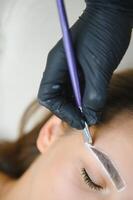 The master holds a white pencil and draws the shape of the eyebrows for permanent makeup photo