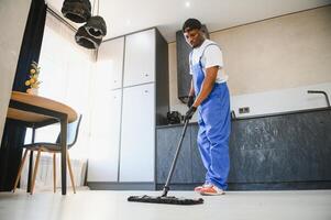 Professional cleaner in blue uniform washing floor and wiping dust from the furniture in the living room of the apartment. Cleaning service concept photo