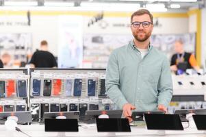 Portrait of happy satisfied young bearded man with eyeglasses testing a tablet in a tech store photo