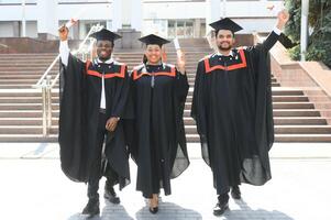 Group of happy smiling multicultural people in graduation gowns and caps with diplomas in hands graduate university outdoors in campus looking at camera. Graduation from college, university concept. photo