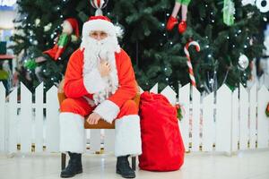 Santa Claus sitting on the background of the Christmas tree in the lobby of the modern shopping mall. Bokeh basic background for design. photo