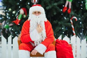 Santa Claus sitting on the background of the Christmas tree in the lobby of the modern shopping mall. Bokeh basic background for design. photo