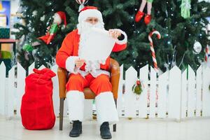 Santa Claus sitting on the background of the Christmas tree in the lobby of the modern shopping mall. Bokeh basic background for design. photo