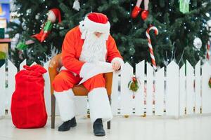 Santa Claus sitting on the background of the Christmas tree in the lobby of the modern shopping mall. Bokeh basic background for design. photo