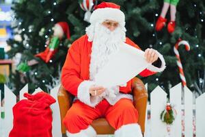 Santa Claus sitting on the background of the Christmas tree in the lobby of the modern shopping mall. Bokeh basic background for design. photo