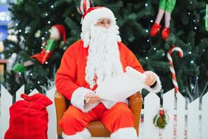 Santa Claus sitting on the background of the Christmas tree in the lobby of the modern shopping mall. Bokeh basic background for design. photo
