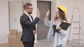 Female designer in helmet with documents giving keys from apartment to young businessman while standing in room video