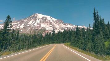 Winding road leading to a snow-capped mountain. A scenic road winds through a lush forest, leading towards a towering snow-capped mountain under a clear blue sky. video