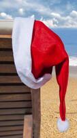 Vertical Santa Hat at the Beach 4K features a beach chair with a Santa hat hanging and gently blowing in the wind with an ocean in the background in a vertical ratio. video