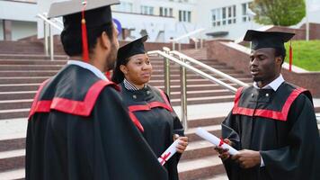 Three multiracial college graduates in graduation gowns talking near university video