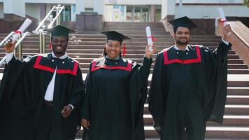 Three college graduates in graduation gowns walking along university campus with diploma video