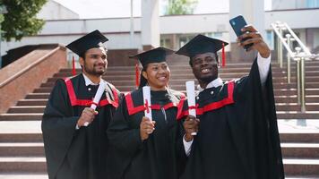 Three happy multiracial diverse students in graduation gowns making selfie and smiling video