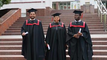 Three college graduates in graduation gowns walking along university campus and talking video