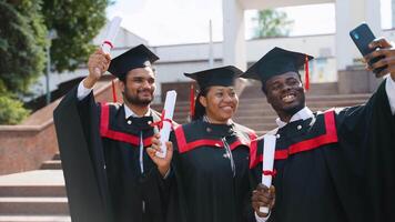 Happy multiracial group of students in graduation dresses and hats taking selfie together video