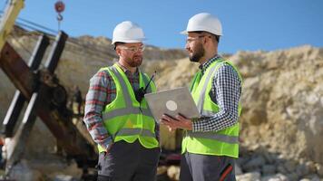Two engineers geologists looks a quarry excavation plan in laptop, excavator in the background video
