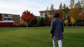 lady walking on a grassy field with autumn trees and buildings in the background video