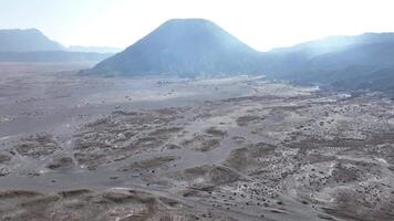 The sweeping aerial view showcases the unique, desert-like terrain, set against the dramatic backdrop of volcanic peaks, creating a surreal and otherworldly atmosphere in East Java. video
