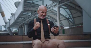 Man sits steps in city, face lit up with joy from what sees screen of mobile phone. Wide smile and animated gestures reveal sincere joy of winning. Man radiates positive energy, rejoicing in victory. video