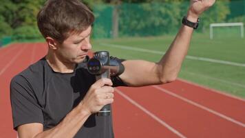 Close-up of male athlete using handheld massage gun on arm muscles for recovery after training session on stadium track. Concept of muscle relaxation and sports recovery video