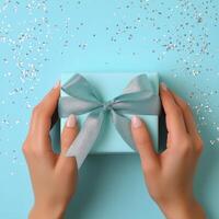 Womans hands holding a wrapped blue gift box with a silver ribbon on a blue background photo