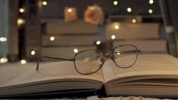 Eyeglasses are resting on an open book. The background features a stack of books, a rose, and warm, twinkling fairy lights video