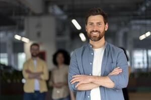 Successful businessman smiling with folded arms in office with coworkers in background photo