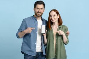Happy couple pointing at smartphone with empty screen on blue background photo