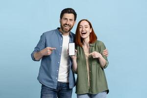 Happy couple pointing at smartphone with empty screen on blue background photo
