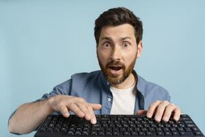 Shocked man looking at camera while typing on keyboard on blue background photo