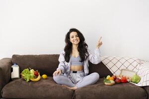 Young woman pointing up and showing healthy food on couch at home photo