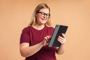 Smiling plus size woman in glasses typing on digital tablet isolated on beige background photo