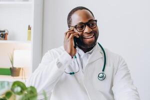 Smiling African American doctor in glasses talking on mobile phone in his office photo