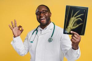 Smiling African American doctor holding X-ray picture with ok gesture, isolated on yellow photo