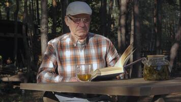 An older gentleman sits at a wooden table, immersed in reading a book. He sips tea from a glass while surrounded by the tranquility of a forest in the afternoon light. video