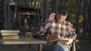 An elderly man sits at a wooden table in a serene forest, absorbed in reading a book while sipping tea from a glass mug, enjoying the tranquility of nature. video