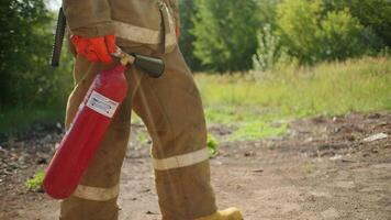 Firefighter with fire extinguisher is walking in woods. Clip. Firefighter epic goes with fire extinguisher in field. Close-up of firefighter carrying fire extinguisher in field on sunny summer day video