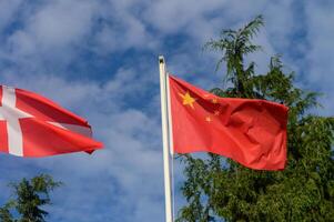 Flags of Denmark and China waving in a gentle breeze against a backdrop of blue skies and lush greenery on a sunny day photo